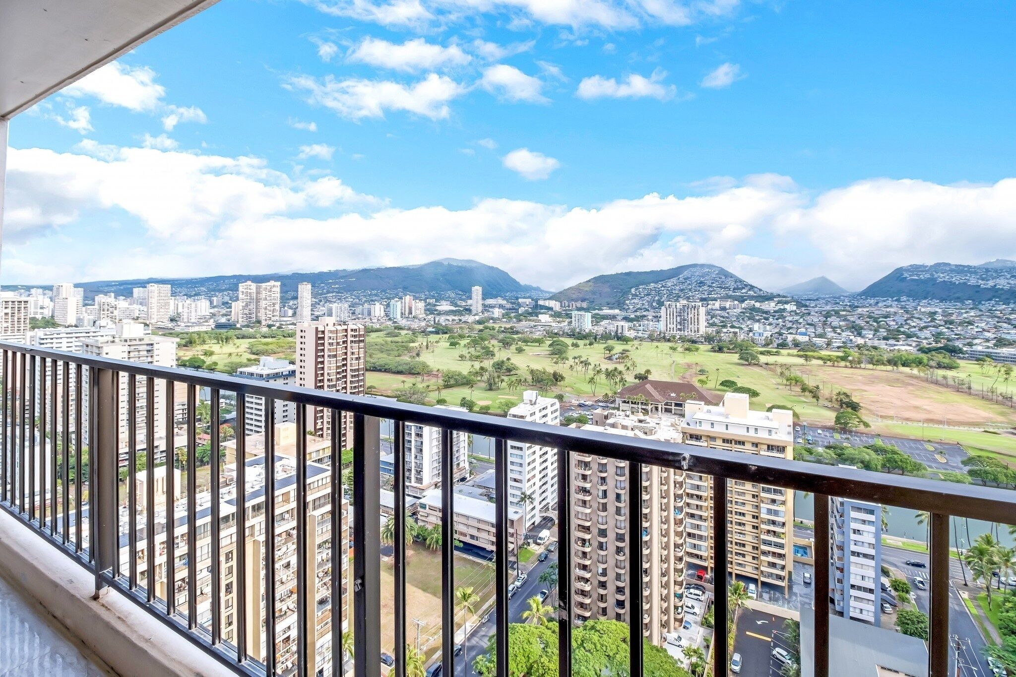 A high-rise building with multiple floors, a balcony with a metal railing, and a cityscape with buildings and greenery in the background.