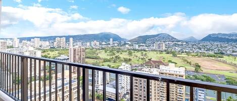 A high-rise building with multiple floors, a balcony with a metal railing, and a cityscape with buildings and greenery in the background.