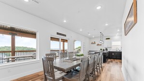 A spacious dining area with a wooden table and chairs, large windows with a view of the outdoors, and a kitchen area with modern appliances and cabinetry.
