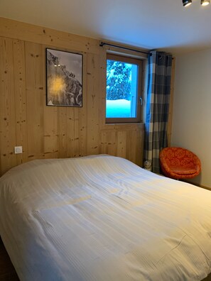 A bedroom with a large bed, wooden walls, a window with a view of snow-covered trees, and a red chair.