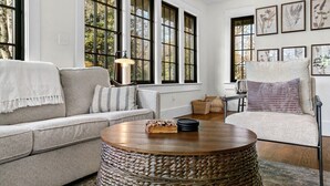 A cozy living room with a grey sofa, a round wooden coffee table, and a chair with a cushion. Large windows allow natural light to fill the space.