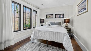 A bedroom with a large bed, two bedside tables, a rug, and a view of trees through the windows.
