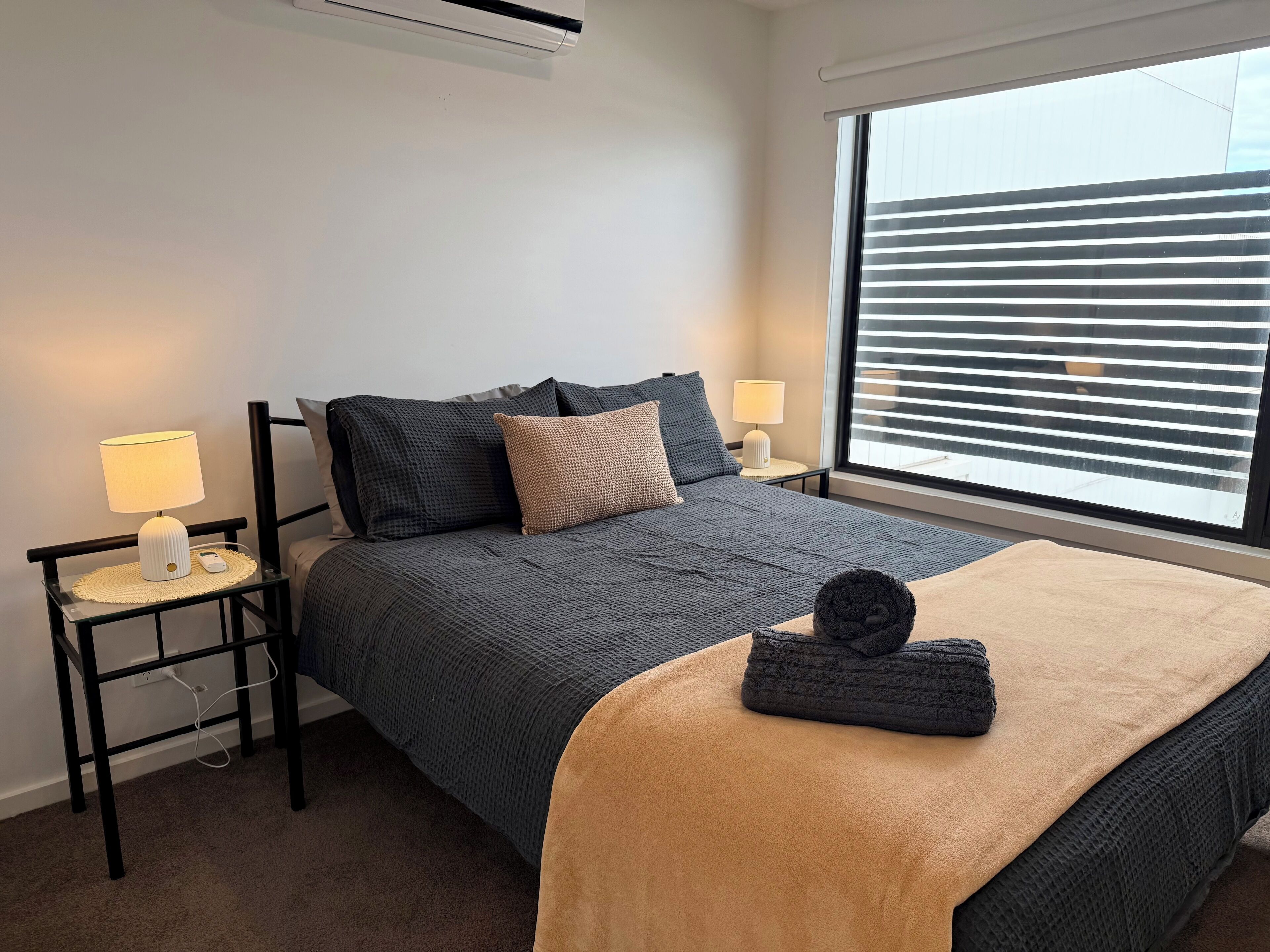 A modern bedroom with a bed, two bedside tables, a lamp, and a window with blinds.