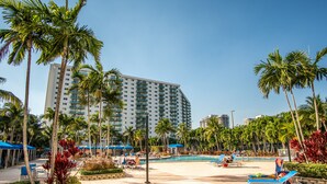 A resort pool area with palm trees, lounge chairs, and a large building in the background.