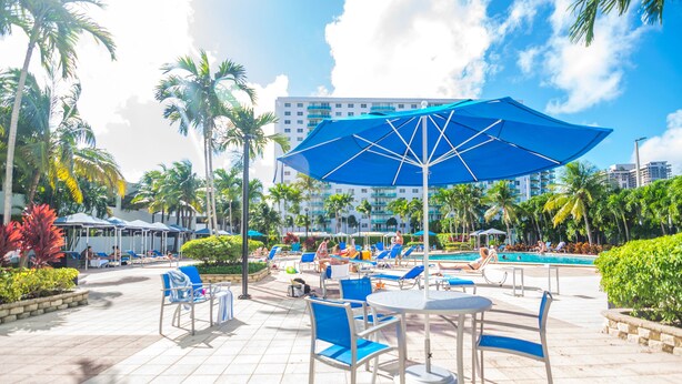 A resort pool area with lounge chairs, umbrellas, and a large blue umbrella providing shade.