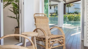 A wicker chair with a cushioned seat and backrest, positioned near a glass door leading to an outdoor area with a view of greenery and a pool. - Abode La Vie (kingscliff)