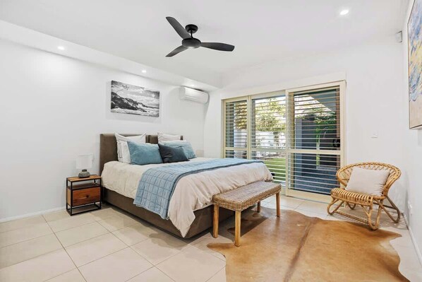 A bedroom with a bed, a ceiling fan, a window with blinds, a wicker chair, and a rug. - Abode La Vie (kingscliff)