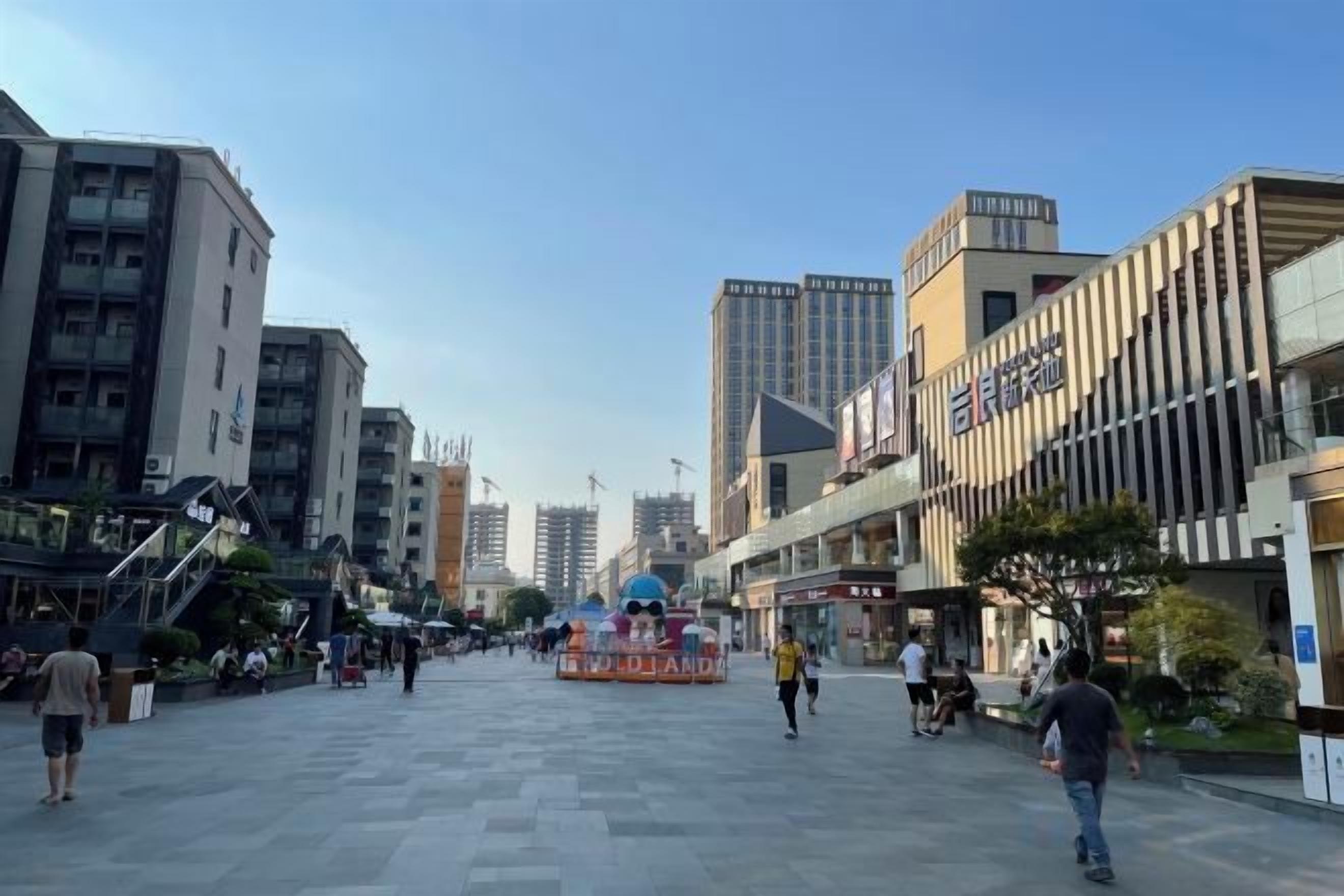 A wide pedestrian street with modern buildings, a clear sky, and a few people walking.