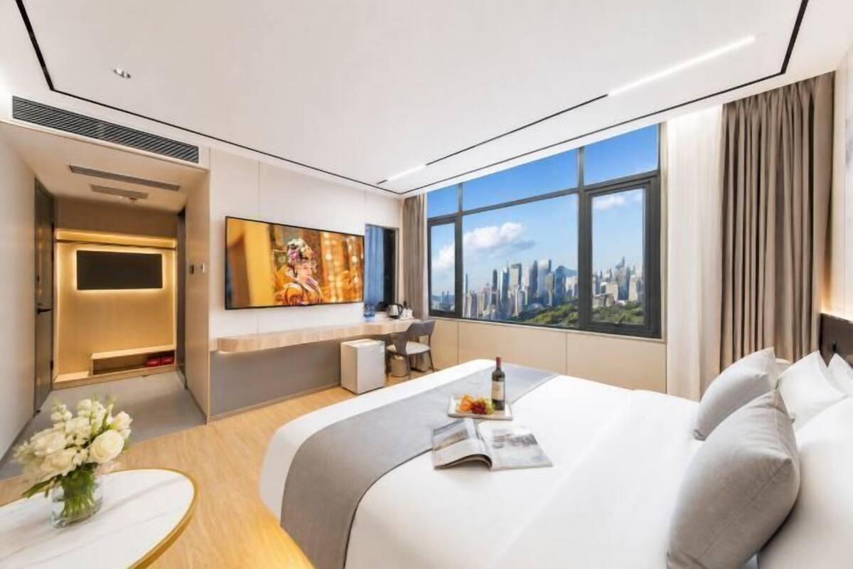 A modern hotel room with a bed, a desk, a TV, and a large window with a city view.