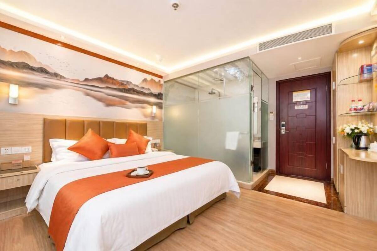 A hotel room with a bed, a glass shower enclosure, a wooden headboard, a wall mural, and a wooden reception desk.