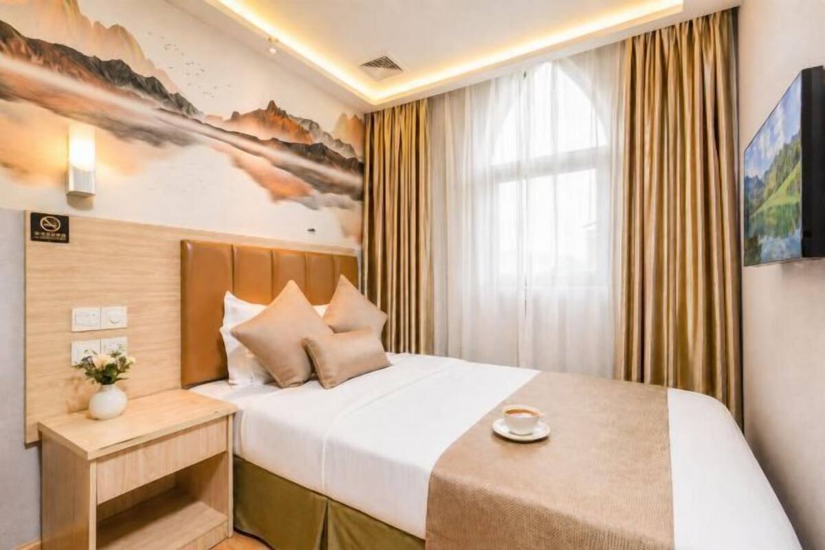 A hotel room with a bed, a nightstand, a wall-mounted TV, and a window with curtains.