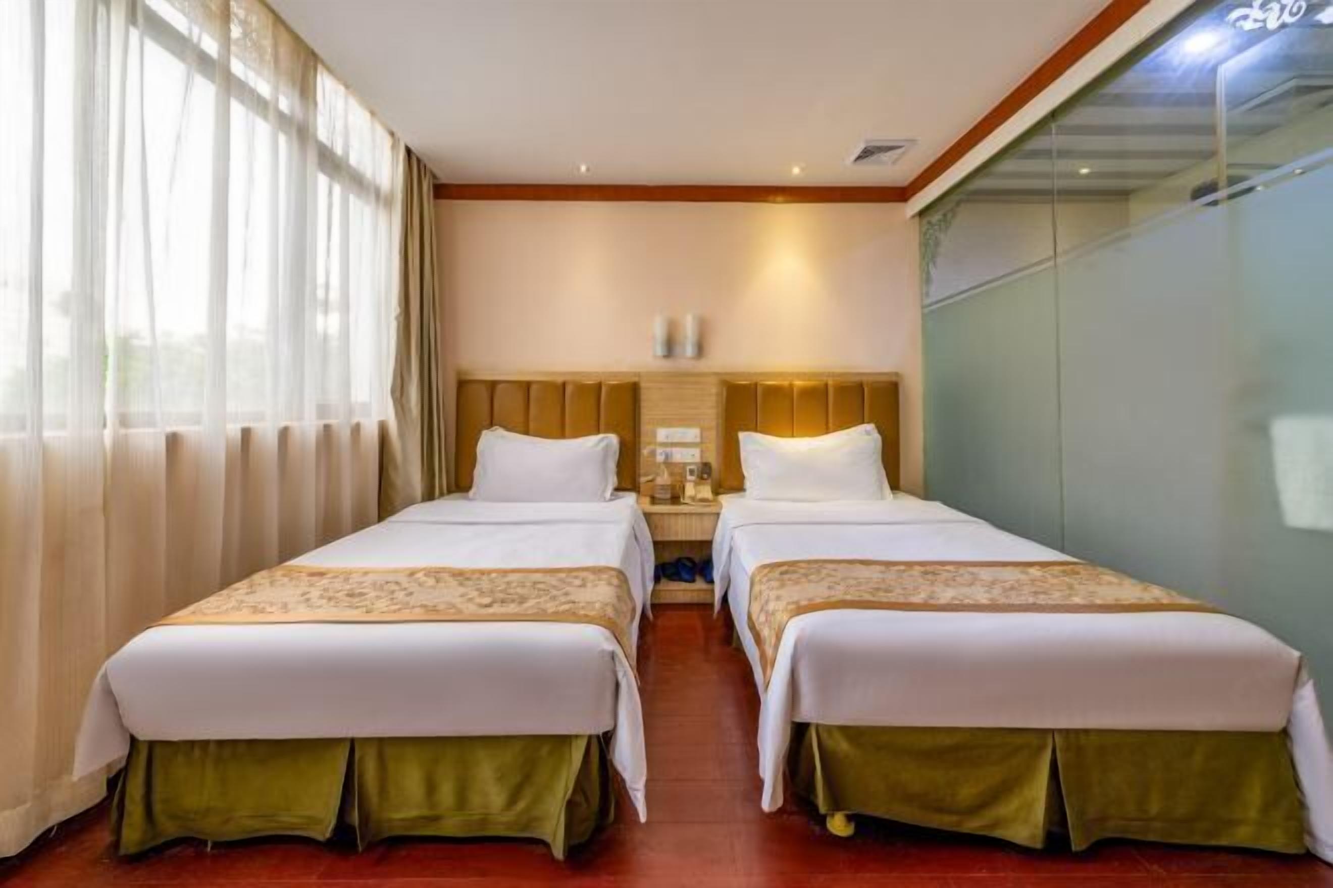 A hotel room with two beds, a nightstand, a lamp, and a view of the outdoors through large windows.
