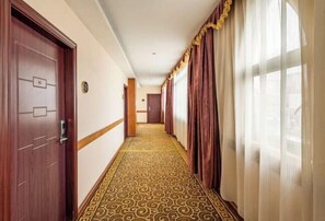 A hotel hallway with patterned carpet, wooden doors, and a view of the outside through sheer curtains. - Forbes Hotel (Shenzhen Longhua Dalang Commercial Center) (Shenzhen)