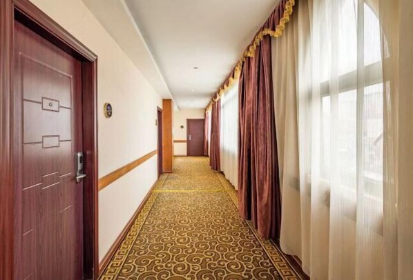 A hotel hallway with patterned carpet, wooden doors, and a view of the outside through sheer curtains. - Forbes Hotel (Shenzhen Longhua Dalang Commercial Center) (Shenzhen)