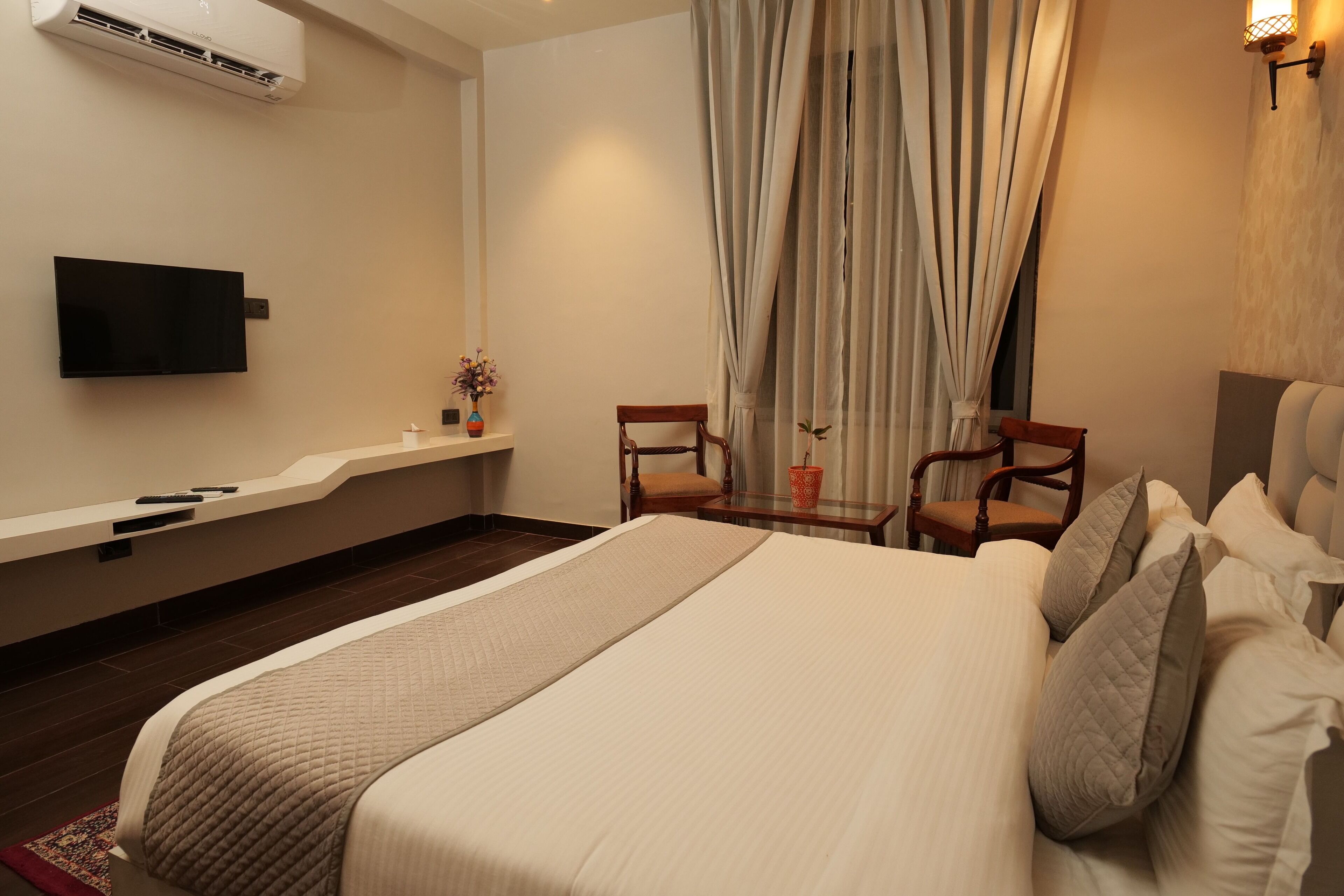 A hotel room with a large bed, a flat-screen TV mounted on the wall, a small table with a vase of flowers, and a chair.