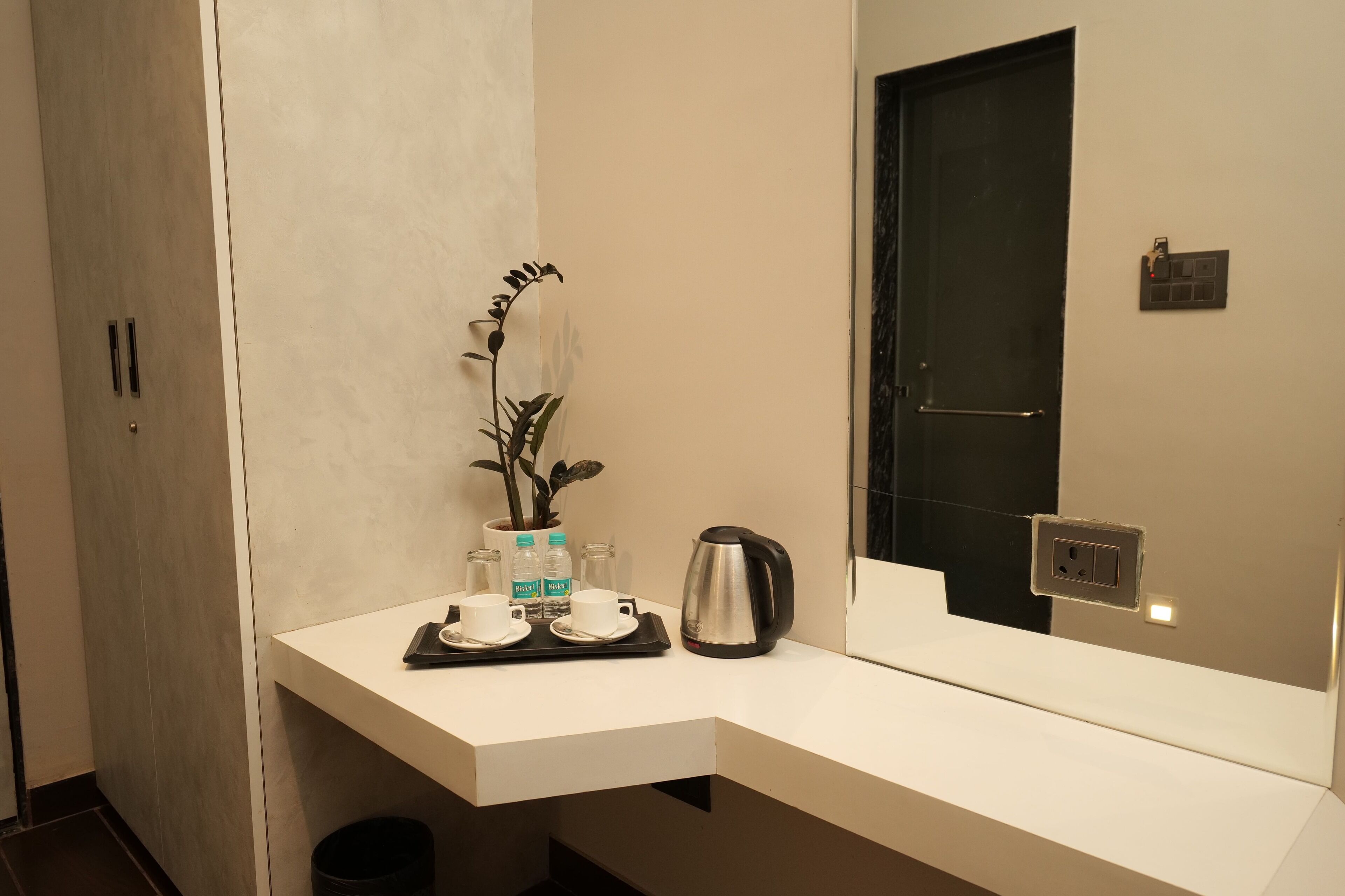 A modern bathroom with a white countertop, a potted plant, two water bottles, a kettle, and a mirror reflecting the door.