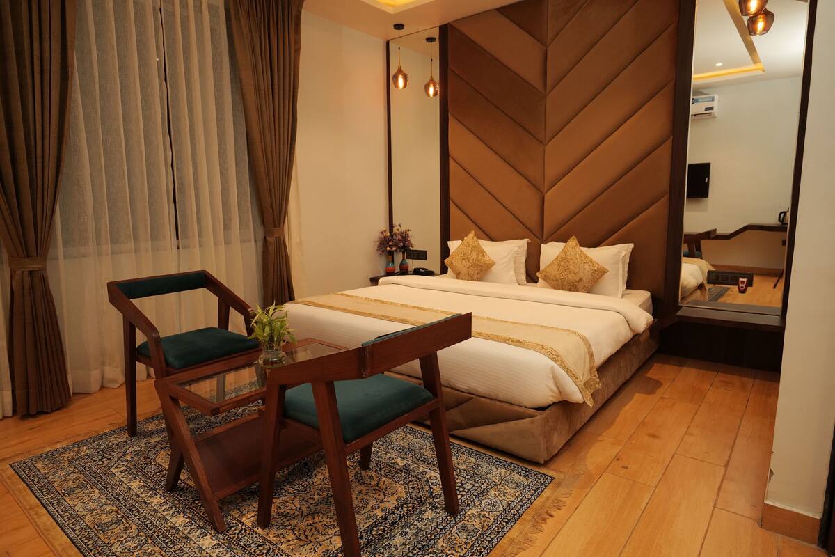 A hotel room with a bed, a wooden headboard, a nightstand, a mirror, a desk with a chair, and a patterned rug.