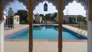 A swimming pool with a view through an ornate archway, surrounded by a tiled deck and a small building in the background. - Hotel 777, Pushkar (Pushkar)