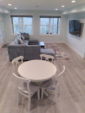 A modern living room with a grey sectional sofa, a white round table with chairs, and large windows with blinds.