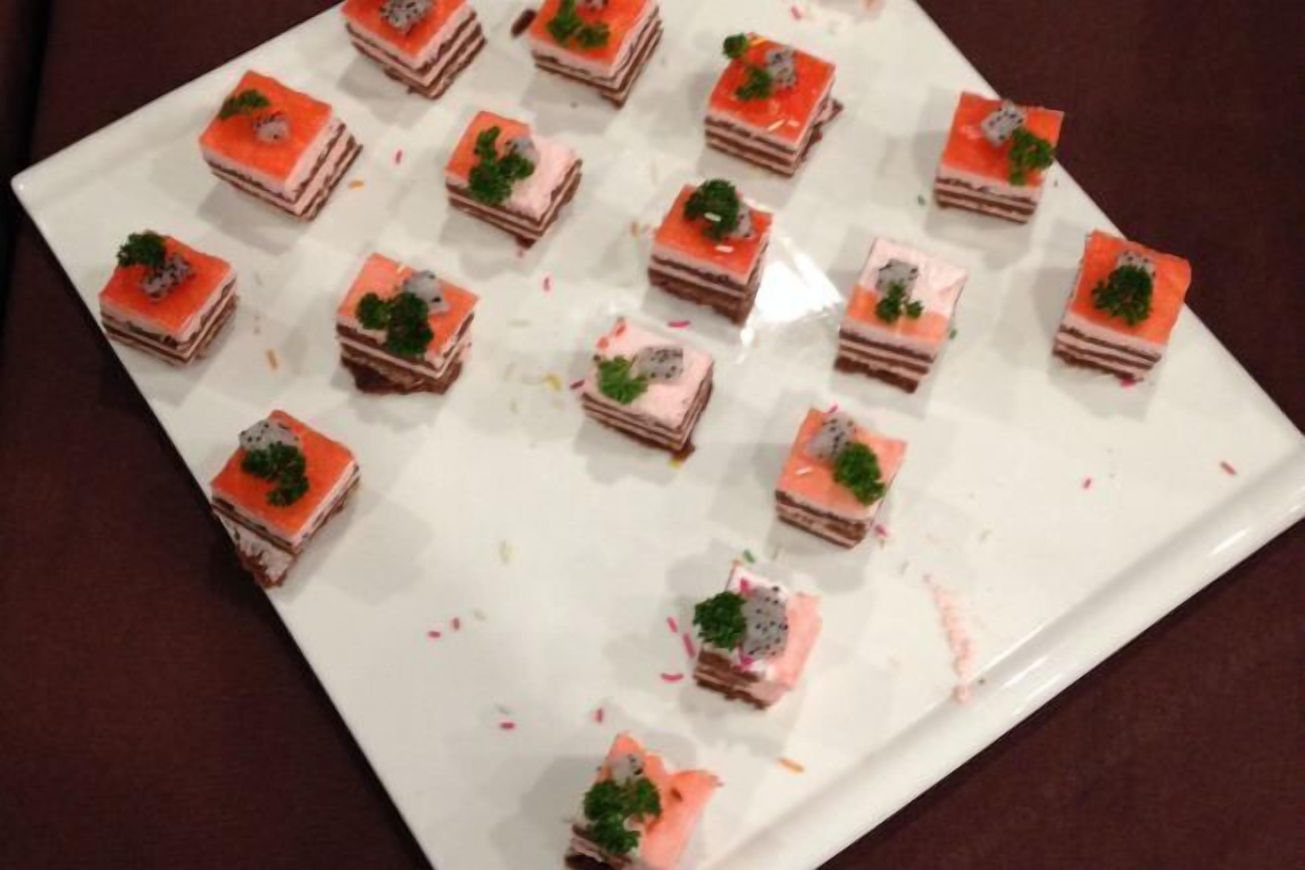 A white rectangular plate with neatly arranged square pastries, each topped with a dollop of pink cream and a small green garnish.