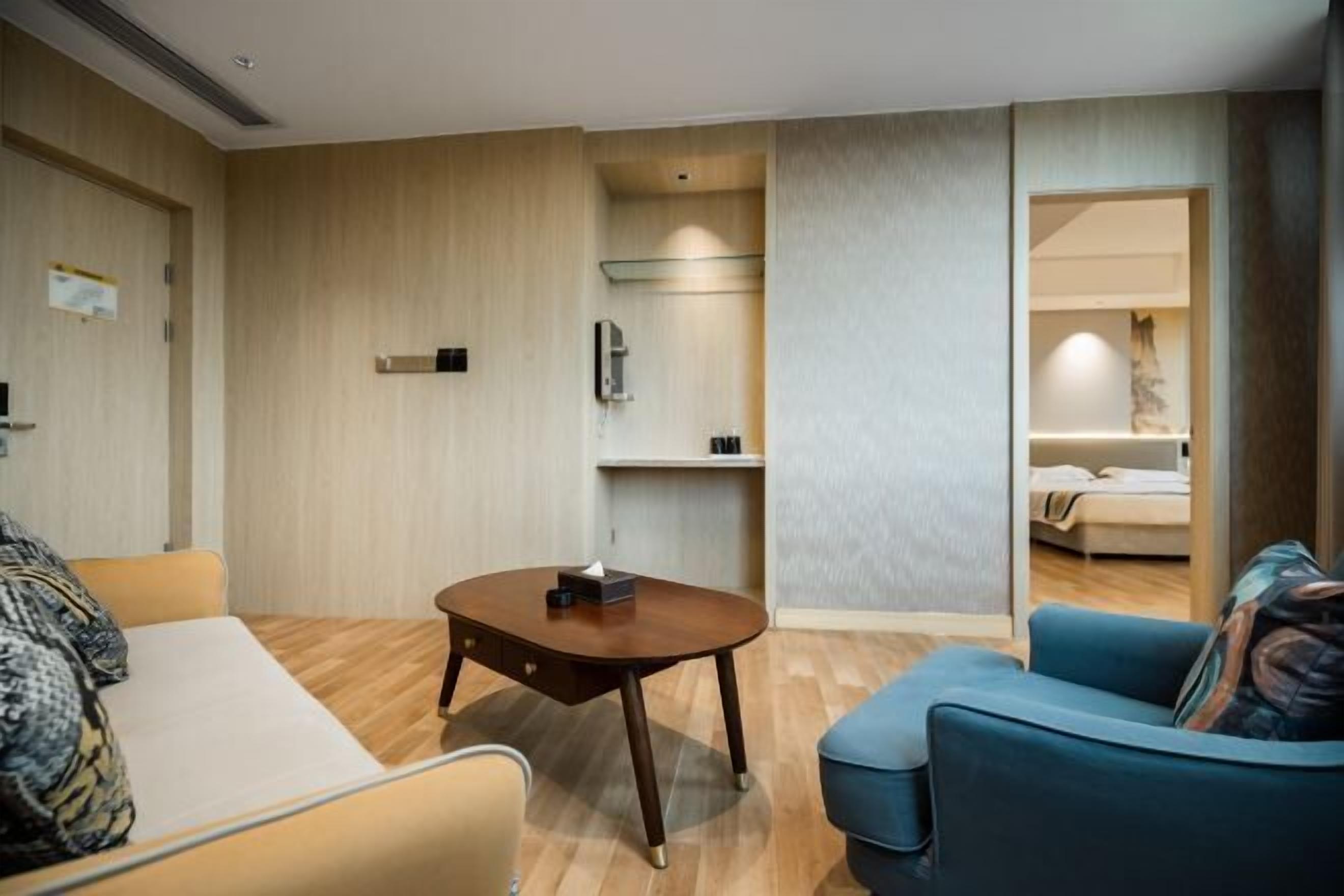 A modern hotel room with a wooden floor, a sofa, a blue armchair, a small table, and a bed in the background.