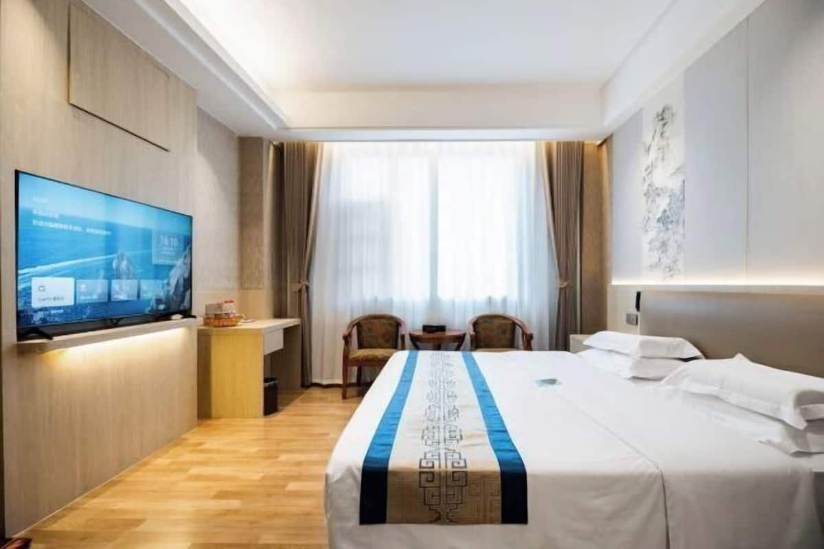 A hotel room with a bed, a TV mounted on the wall, a small table, two chairs, and a window with curtains.