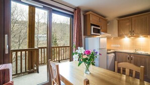 A dining area with a wooden table and chairs, a vase with flowers, and a view of the outdoors through large sliding glass doors. - Wifi, swimming pool, terrace, balcony, parking, tv, ski locker, 24m², Les Bottières (Saint-Pancrace)
