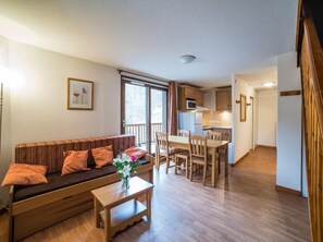 Ein gemütlicher Wohnbereich mit einer Couch, einem Couchtisch und einem Esstisch mit Stühlen. Im Hintergrund ist eine Küchenzeile mit Kühlschrank und Schränken zu sehen.
