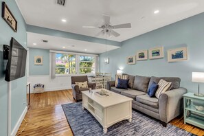 A living room with a grey sofa, a white coffee table, and a ceiling fan. There are framed pictures on the wall and a window with a view of greenery. - Dog friendly cottage - One block to the Bay! (Anna Maria)