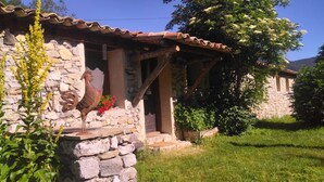 A stone building with a rustic roof, a rooster statue, and a garden with grass and flowers. - "La Bergerie "Gîte at the end of the world. La Valbonnette (Barrême)