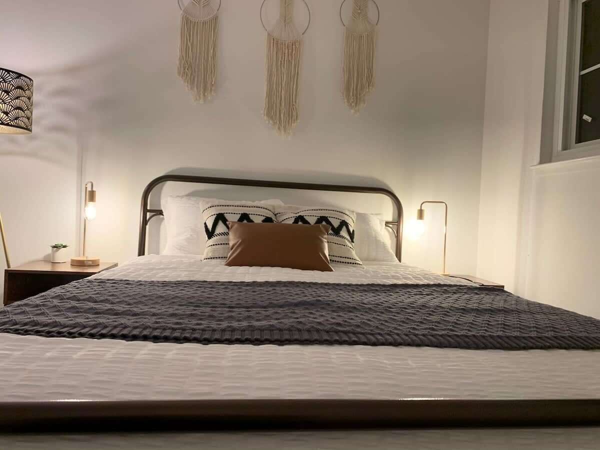 A neatly made bed with a patterned blanket, a single brown pillow, and a headboard with decorative tassels. There are bedside lamps and a small table with a plant.