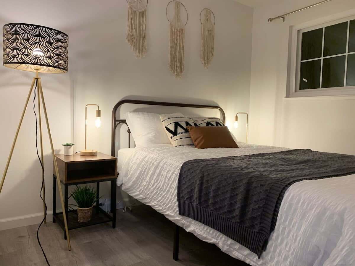 Un llit ben arropat amb una manta fosca, una taula de nit amb una làmpada i penjolls de macramé fixats a la paret.