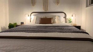 Una cama bien tendida con un cabecero oscuro, una mesita de noche con una lámpara y adornos de macramé en la pared.