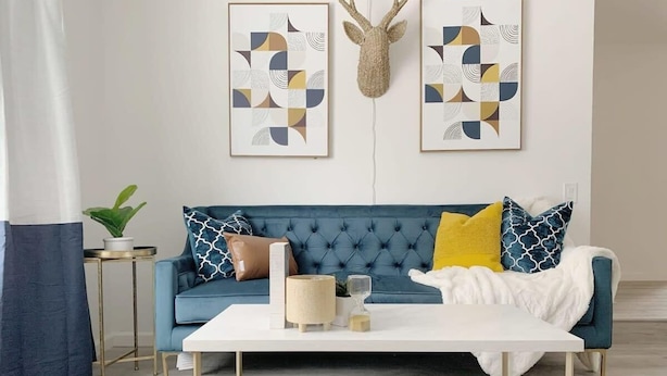 Una sala de estar con un sofá azul, una mesa de centro blanca y un tapiz de piel de vaca. Hay dos cuadros abstractos enmarcados en la pared y una cabeza de venado montada sobre el sofá.