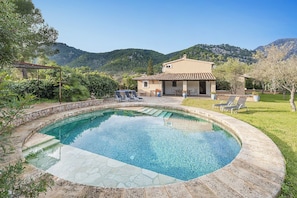 Uma piscina com borda de pedra, um alpendre com espreguiçadeiras e uma casa com telhado em cerâmica.