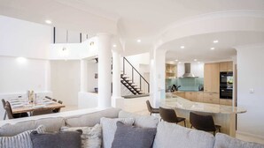 Un vaste espace de vie doté d’un haut plafond, une table à manger pour six personnes, un canapé agrémenté de nombreux coussins, une cuisine équipée d’appareils modernes et un escalier menant à un niveau supérieur.