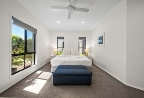A bedroom with a large window, a bed, a nightstand, a bedside table, a floor lamp, and a ceiling fan.