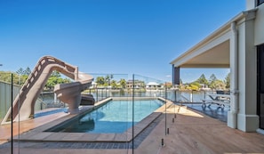 Una piscina con una estructura de tobogán única, rodeada por una valla de cristal, y un área de descanso techada con vista al agua.