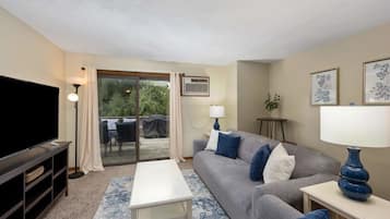 A living room with a flat-screen TV, a grey sofa with blue cushions, a white coffee table, and a blue lamp.