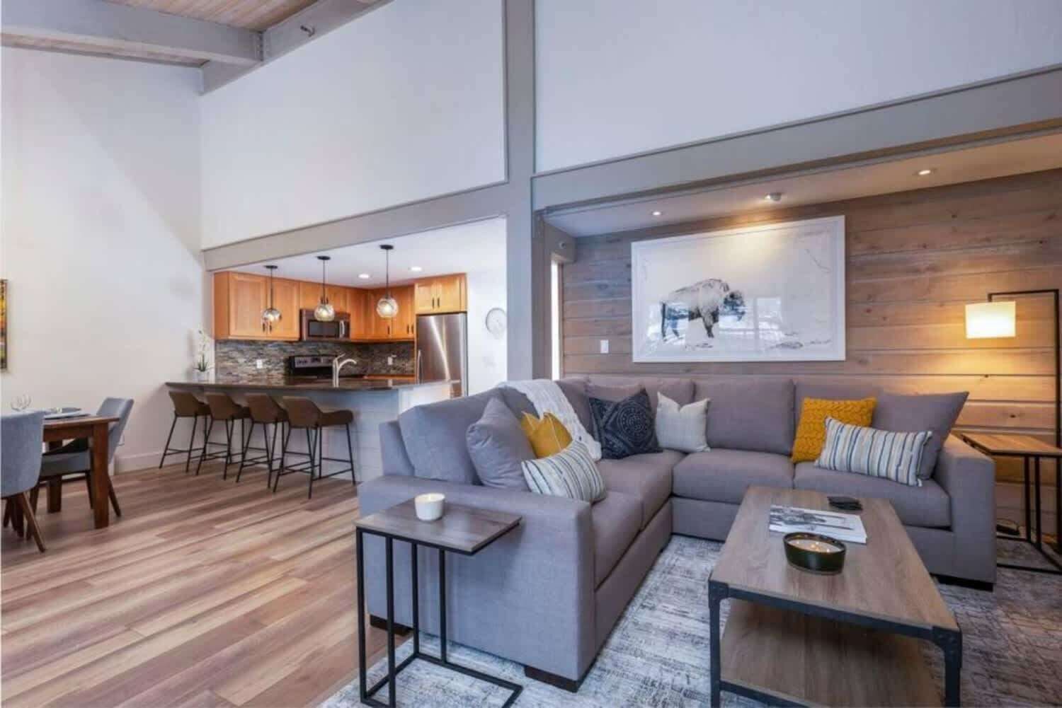 Ein modernes Wohnzimmer mit grauem Sofa, Couchtisch und Barhocker. Es gibt eine Küchenzeile mit Holzschränken und ein großes Bild an der Wand.