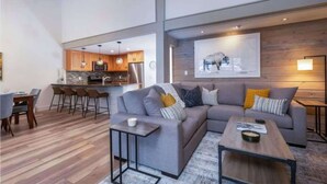 Ein modernes Wohnzimmer mit grauem Sofa, Couchtisch und Barhocker. Es gibt eine Küchenzeile mit Holzschränken und ein großes Bild an der Wand.