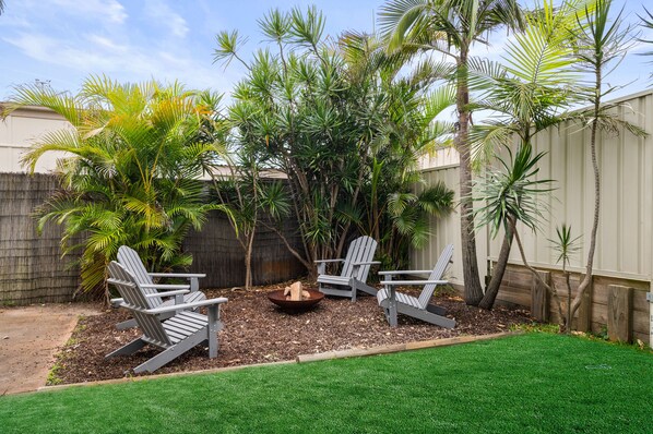 A backyard with a fire pit, two Adirondack chairs, and a variety of tropical plants.