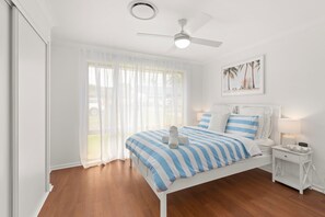 A bedroom with a bed, a nightstand, a ceiling fan, and a window with sheer curtains.