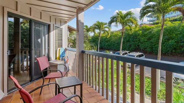 A balcony with a view of a road and parked cars, a table, chairs, and a surfboard.