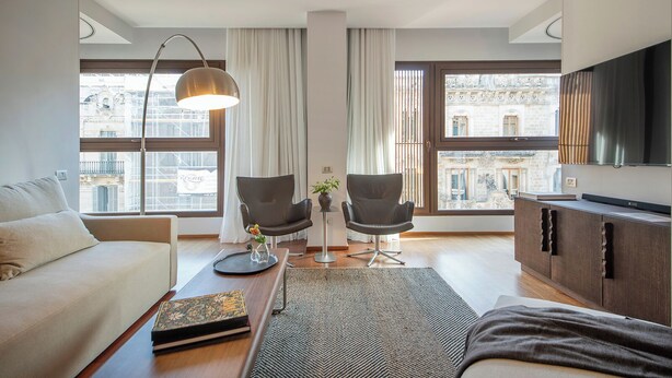 Ein modernes Wohnzimmer mit einer Couch, zwei Sesseln, einem Couchtisch und einem Fernseher. Große Fenster bieten Blick auf die Stadt.