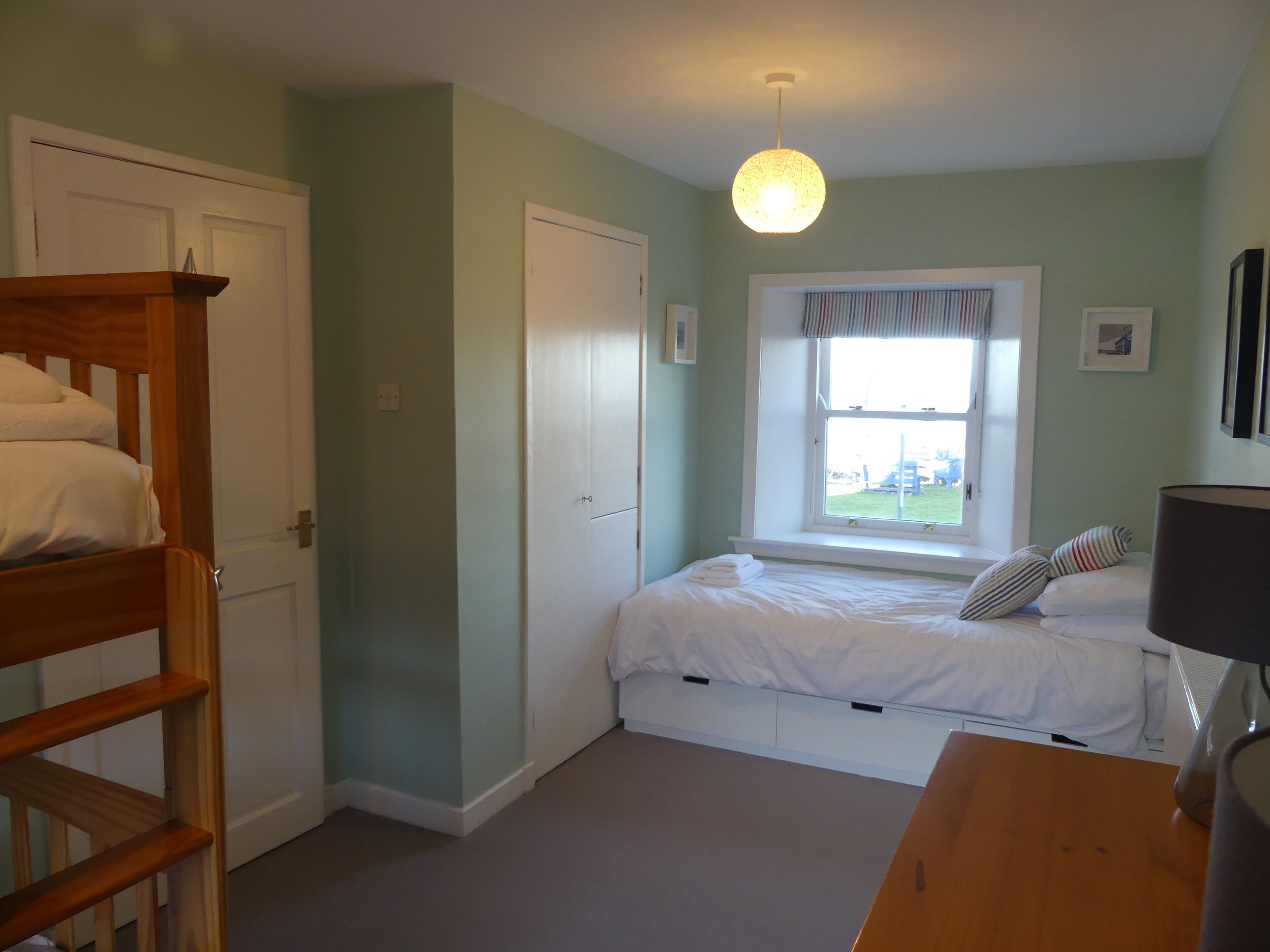 Une chambre avec un lit superposé, un lit simple près d’une fenêtre, un bureau et une lampe.