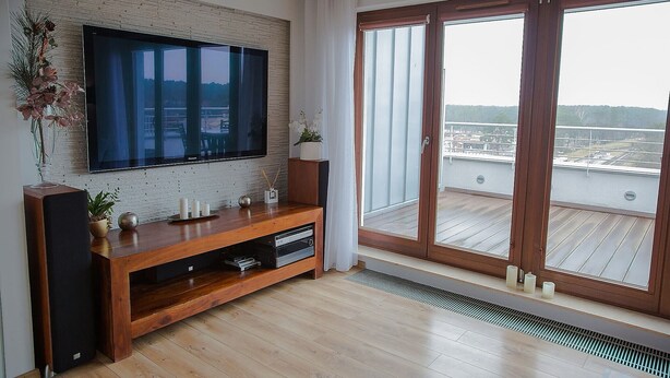 A modern living room with a flat-screen TV, wooden entertainment unit, and a large sliding glass door leading to a balcony with a view of a water body. - Apartamenty Swinoujscie-Wojska Polskiego (Swinoujscie)