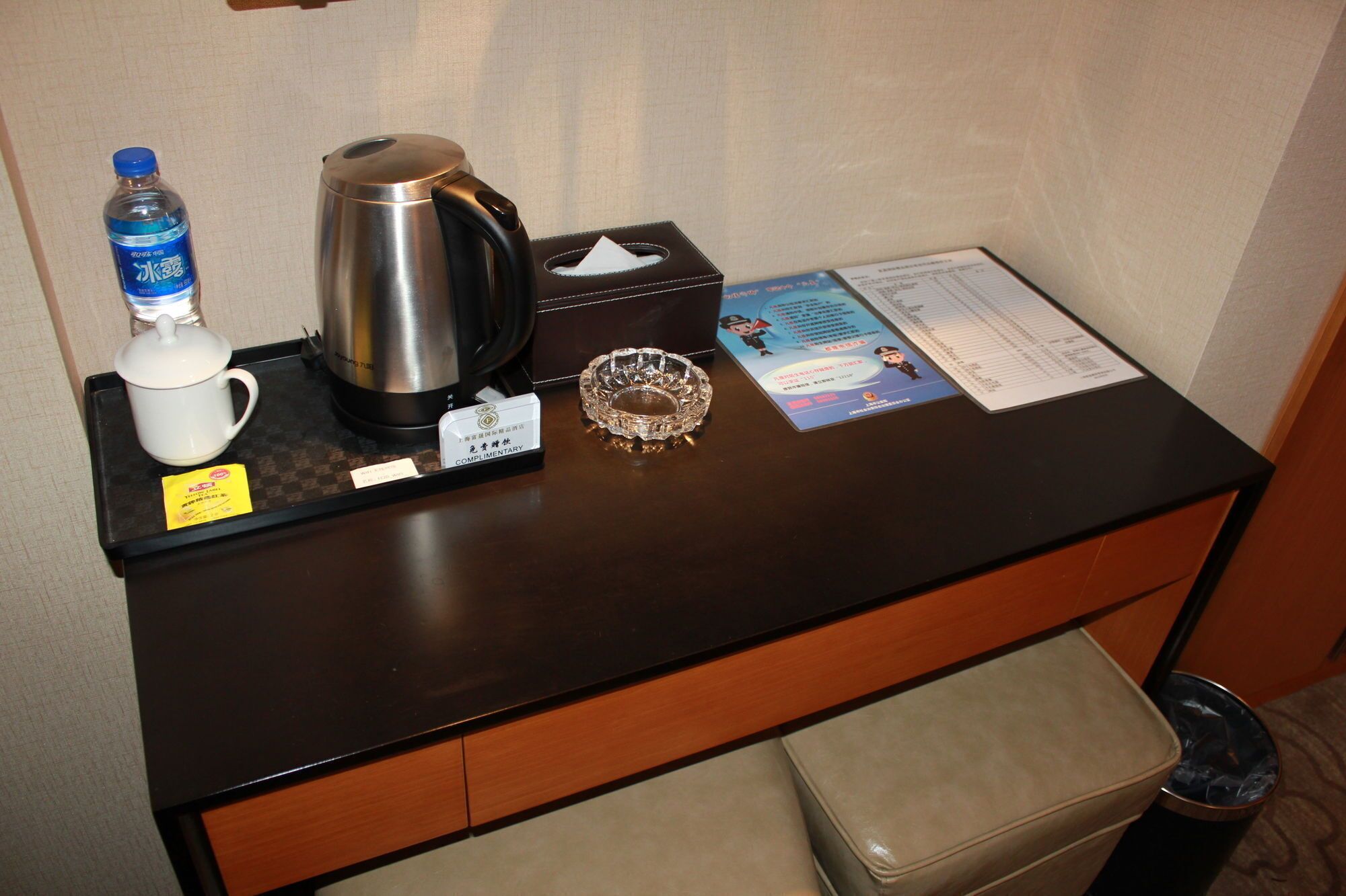 A hotel desk with a water bottle, a coffee pot, a tissue box, a complimentary card, and a menu.