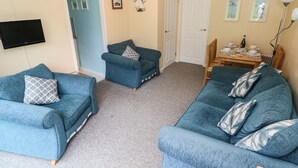 A living room with two blue sofas, a small armchair, a flat-screen TV, a dining table set, and a door with a light switch above it. - Number 6 Oxwich Leisure Park (Swansea)