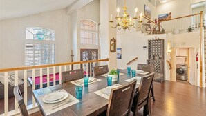 Un coin repas avec une table en bois dressée pour six personnes, entourée de chaises, et un lustre au-dessus.